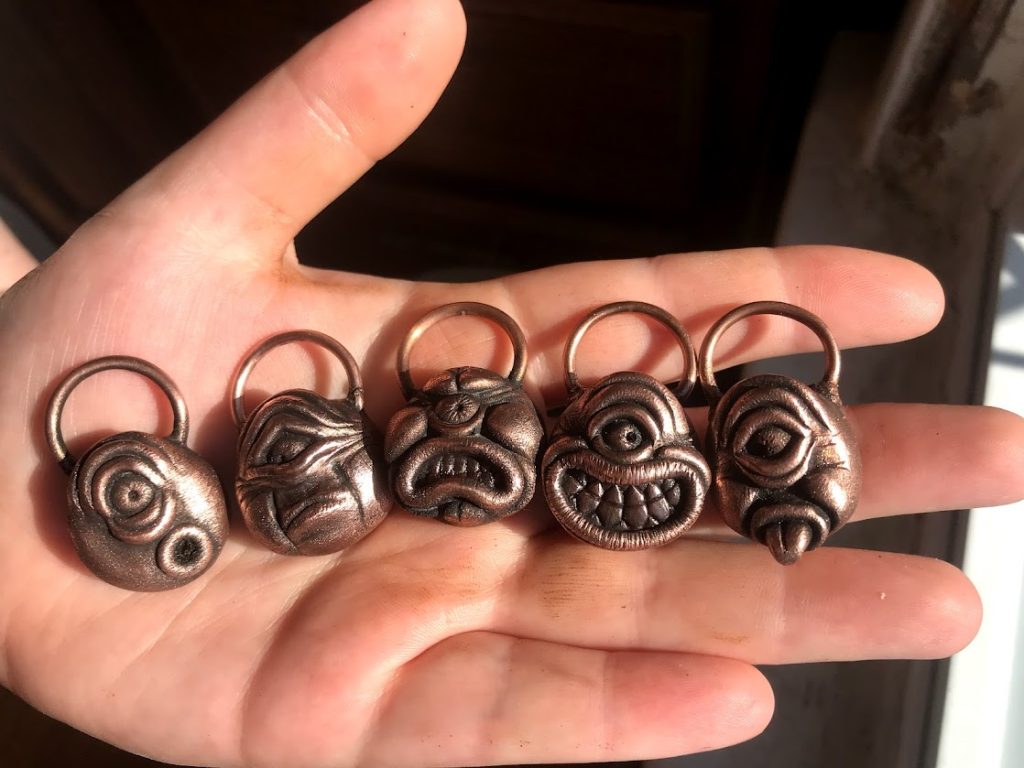Photo of an open palm cradling a set of five copper plated necklace pendants. The pendants are cartoonish faces with expressions ranging from shock to joy.