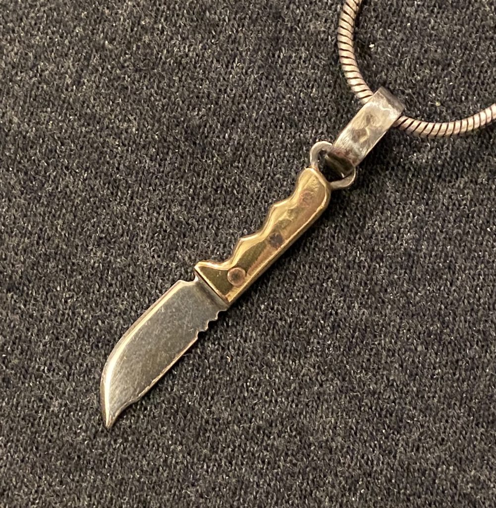 image of a miniature silver and brass fixed blade knife pendant set against a plain gray backdrop.