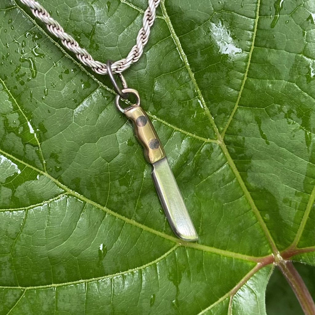 Image of a miniature silver and brass machete pendant set against a green leaf background.