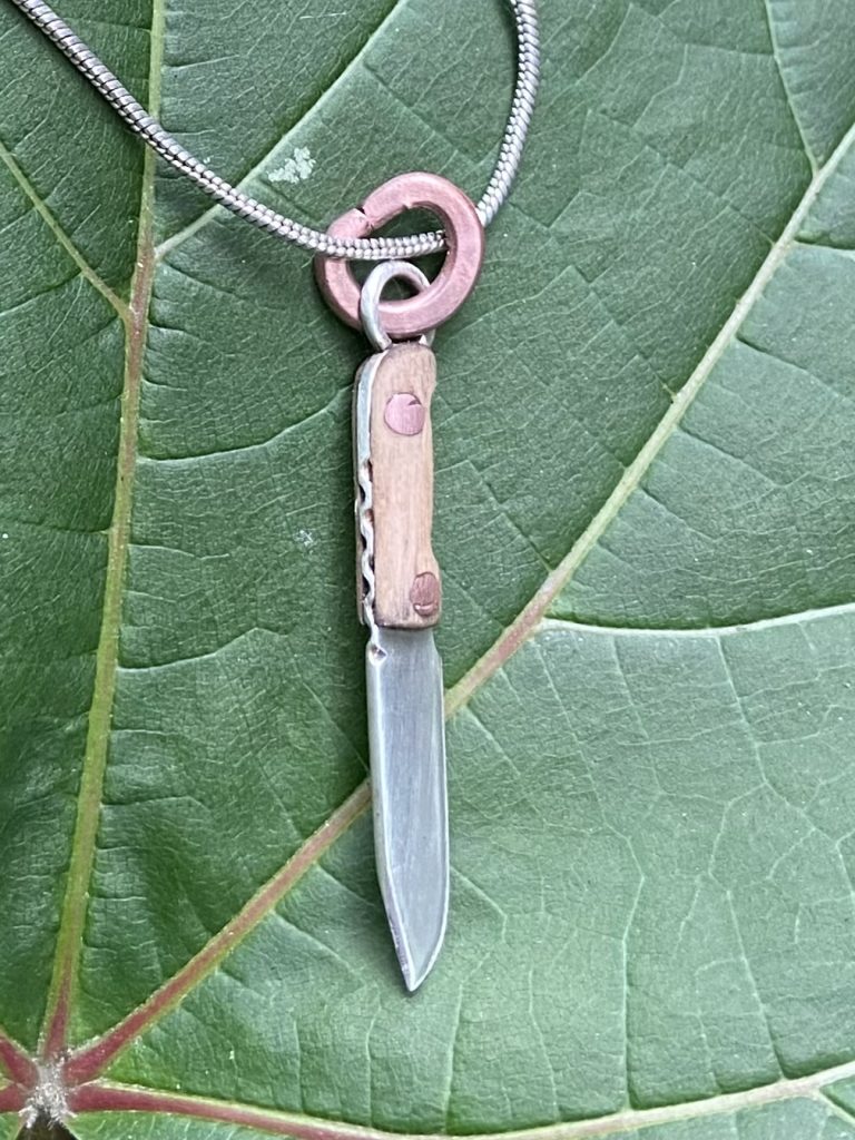 Image of a fabricated miniature clip-point knife pendant set against a green-leaf background.