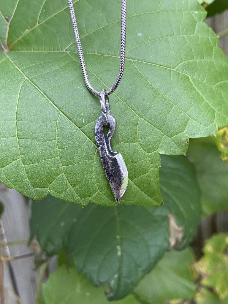 Image of a silver fabricated minature curved tang knife set against a green-leaf background.